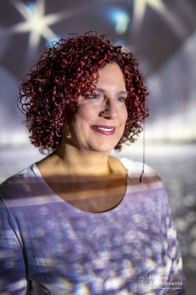 Frau mit lockigem, rotem Haar lächelt vor einem funkelnden, verschwommenen Hintergrund.