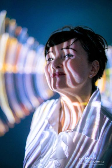 Frau mit gestreiften Lichtmustern auf ihrem Gesicht vor einem bunten Hintergrund.