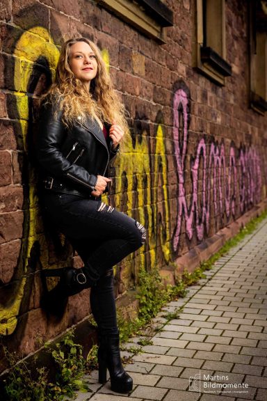 Frau mit lockigem Haar in Lederjacke und Jeans, posiert vor einer bunten Graffiti-Wand.