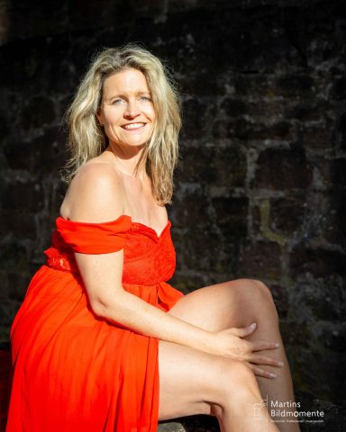 Frau in rotem Kleid sitzt lächelnd vor einer düsteren Steinmauer.